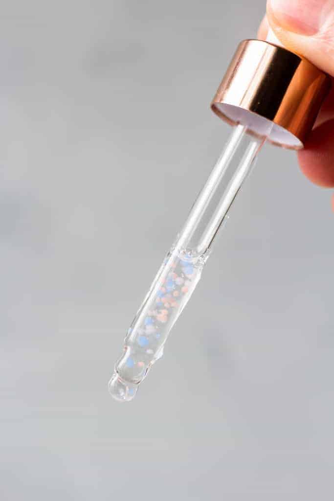 A clear dropper filled with a clear serum with pink and blue molecules in it
