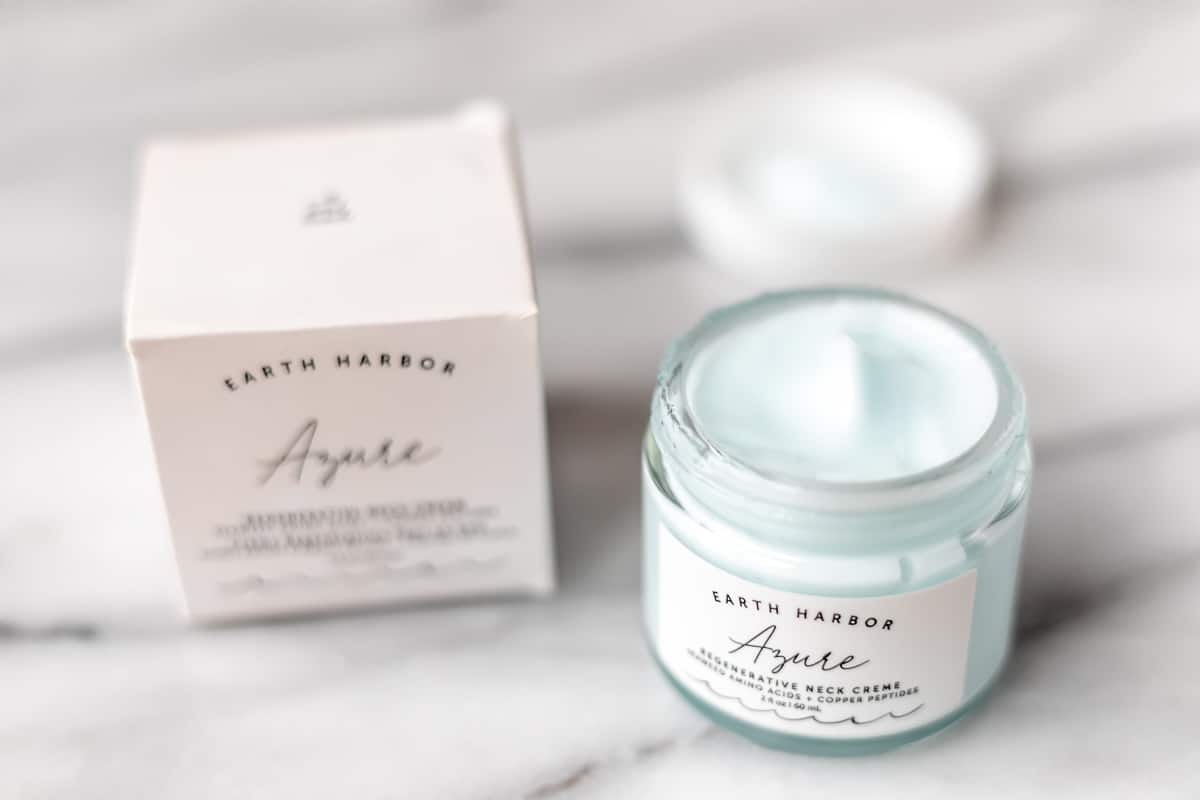 An opened jar of Earth Harbor neck cream next to it's box and lid on a marble background.