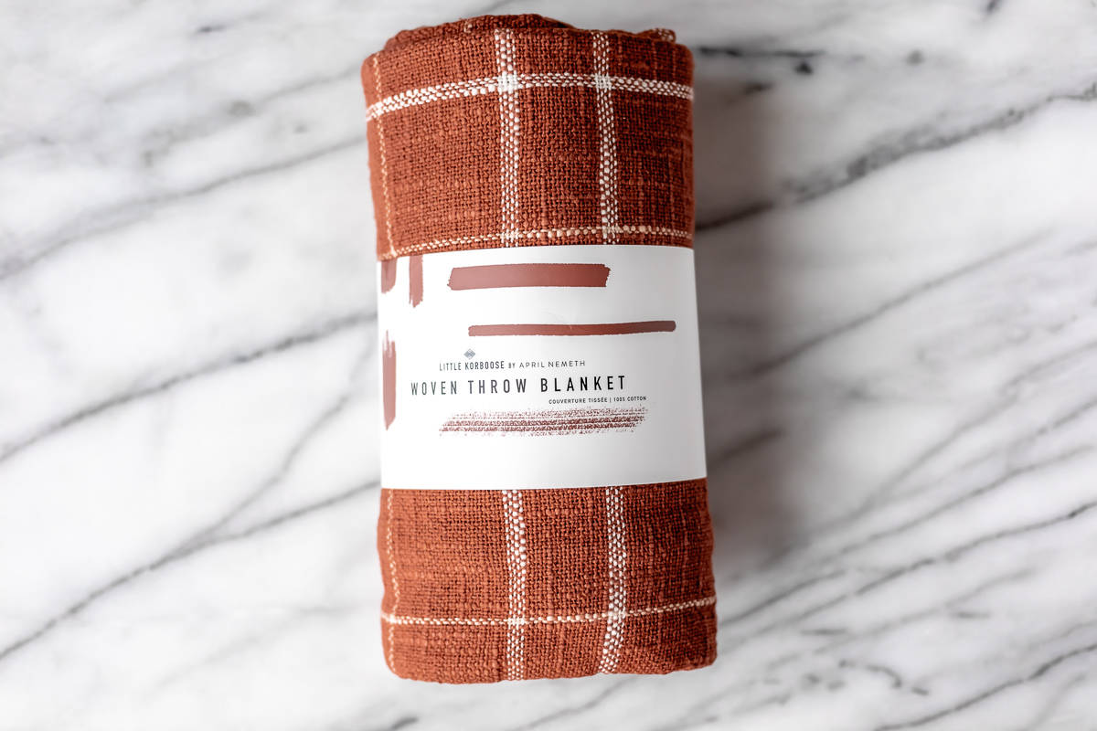 A rust color woven blanket rolled up in it's packaging on a marble background.