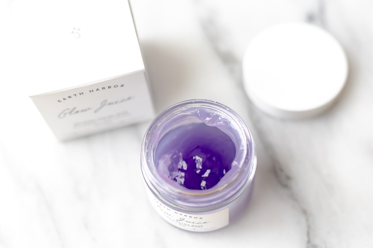 An opened jar of Earth Harbor Glow Juice mask which is a purple jelly-like substance with the lid and box in the background on a light colored surface.