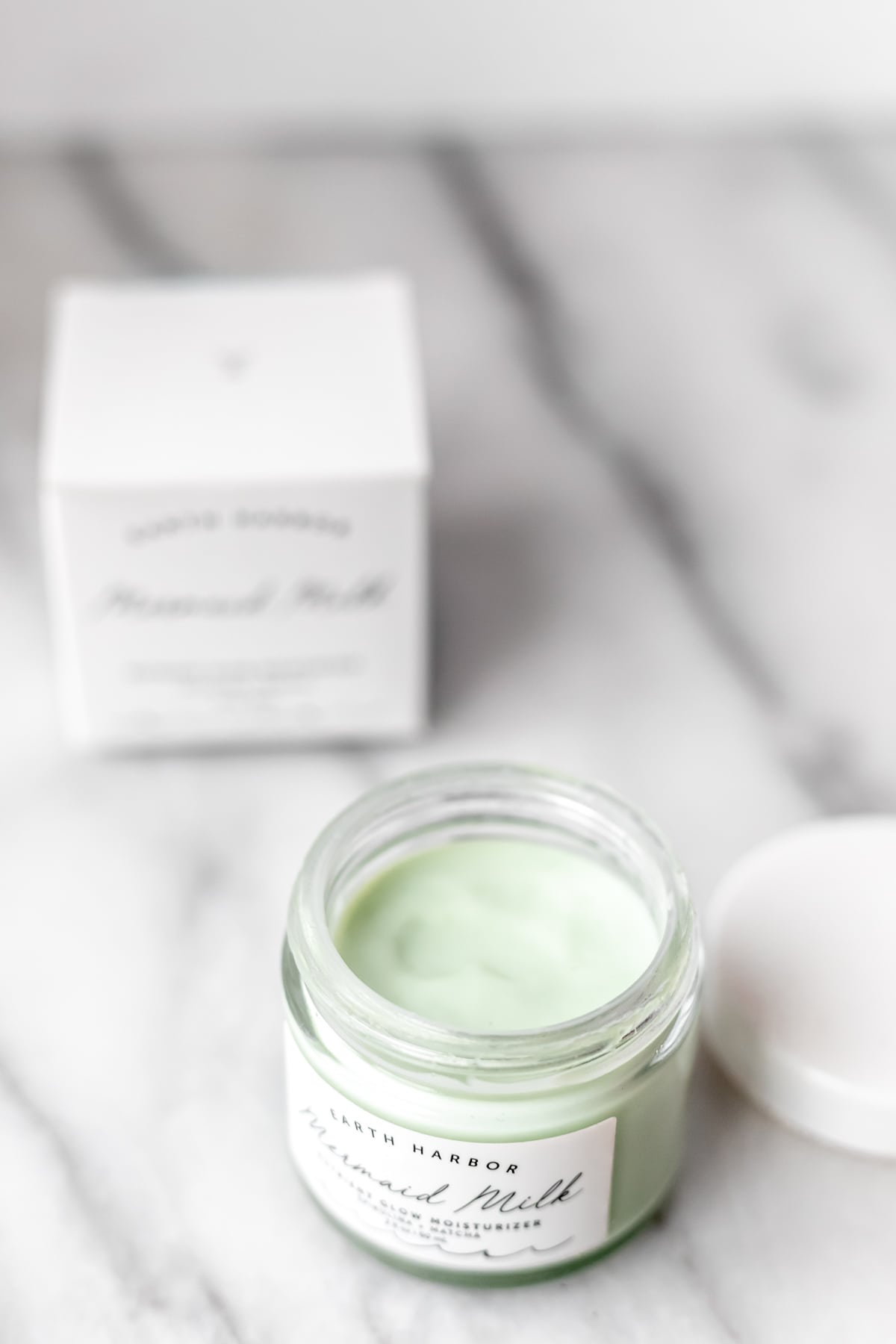 An opened jar of Earth Harbor Mermaid Milk showing the light green cream inside with the lid and box around it.