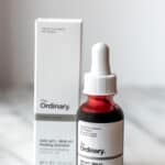 A bottle of The Ordinary Peeling Solution in front of the box it comes in on a marble table with a light background.