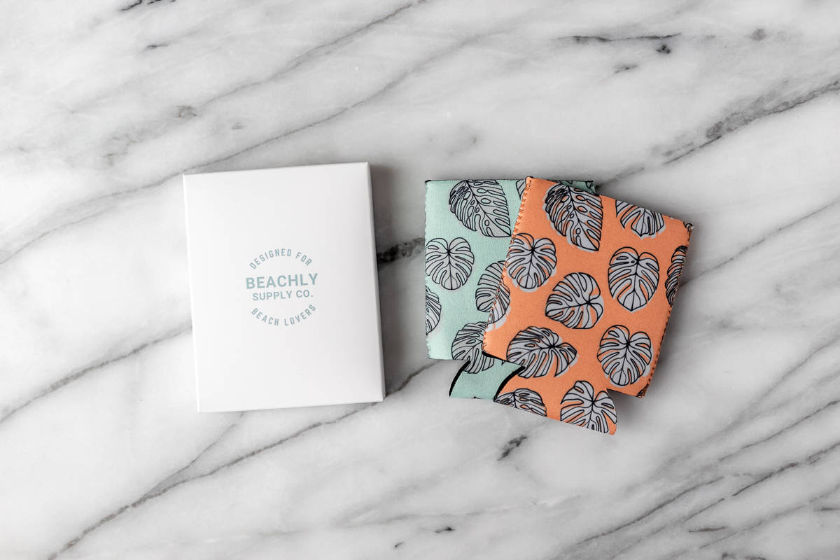 Two ‘Cheers’ Can Cooler Pack by Beachly in orange and teal with monstera leaves on them with the box next to them.