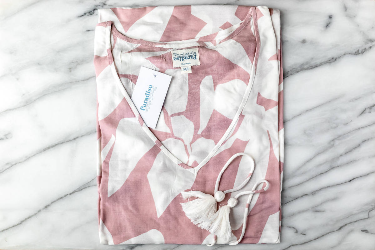The Islander Coverup Dress by Poolside Paradiso in pink and white folded on a marble background.